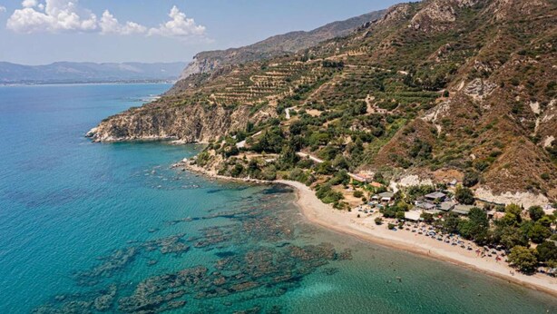 Une zone côtière caractérisée par des eaux turquoise et limpides, un terrain rocheux et une plage bordée de bâtiments et de végétation.