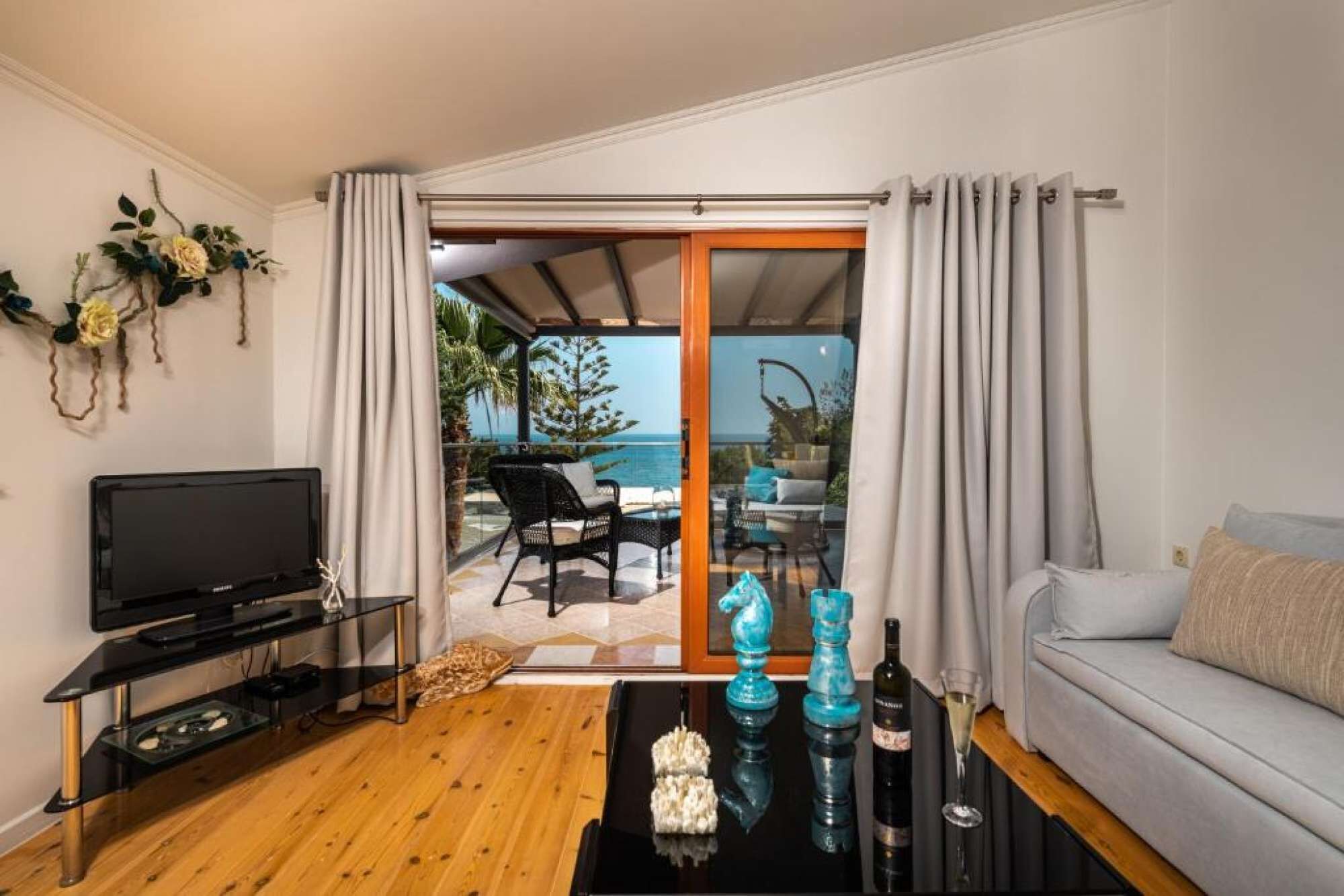 A living room with a TV, a sofa, a coffee table with decorative items, and a view of the sea through a glass door.