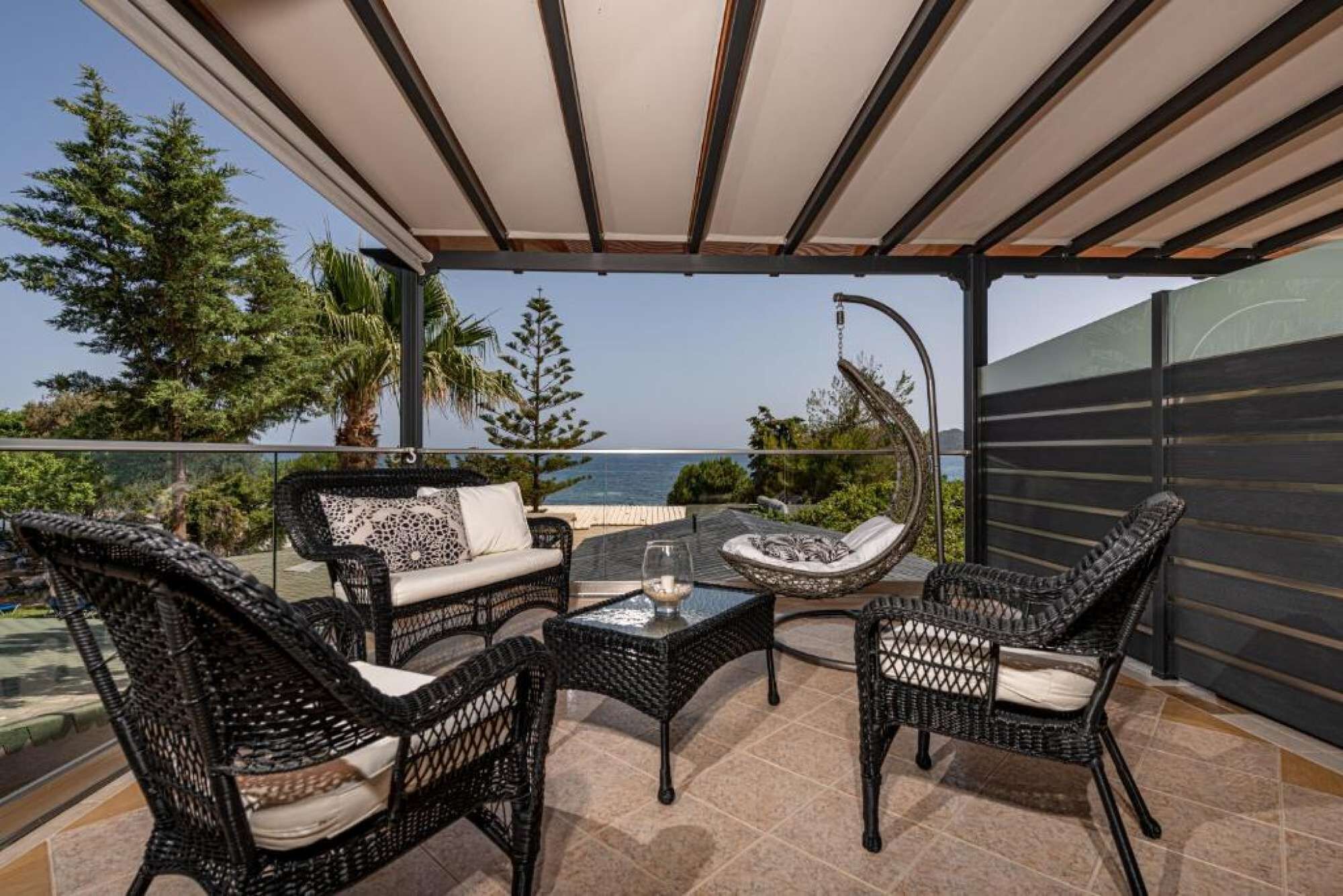 A covered outdoor seating area with wicker furniture, a glass table, and a view of the sea.