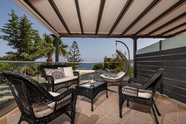 A covered outdoor seating area with wicker furniture, a glass table, and a view of the sea.