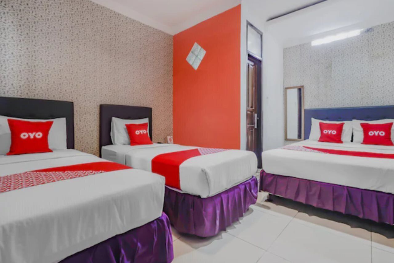 A hotel room with three beds, each with a red 'OYO' pillow, white bedding, and purple skirts.