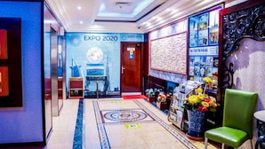 A hotel lobby with a blue wall featuring the Expo 2020 logo, a reception desk, a green chair, and a wall with various photos and a clock. - Phoenix Hotel Deira (Dubai)