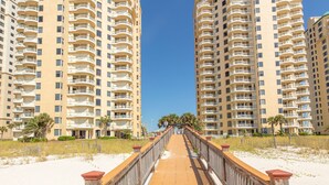 A wooden boardwalk leading to two high-rise buildings with balconies, surrounded by sand and sparse vegetation. - Breathtaking Panoramic Views of the Gulf (Pensacola)
