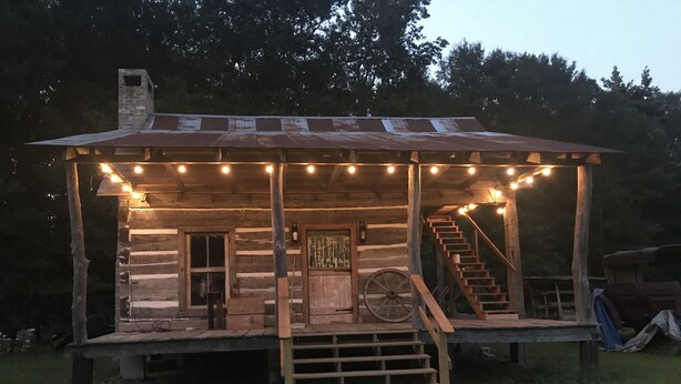 A rustic wooden cabin with a porch, a wooden staircase, and a wheelbarrow. - 1830’s Log Cabin (Franklinton)