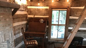 A rustic cabin interior with a wooden dining table, a chair, a small rug, and a window with a view of trees. - 1830’s Log Cabin (Franklinton)