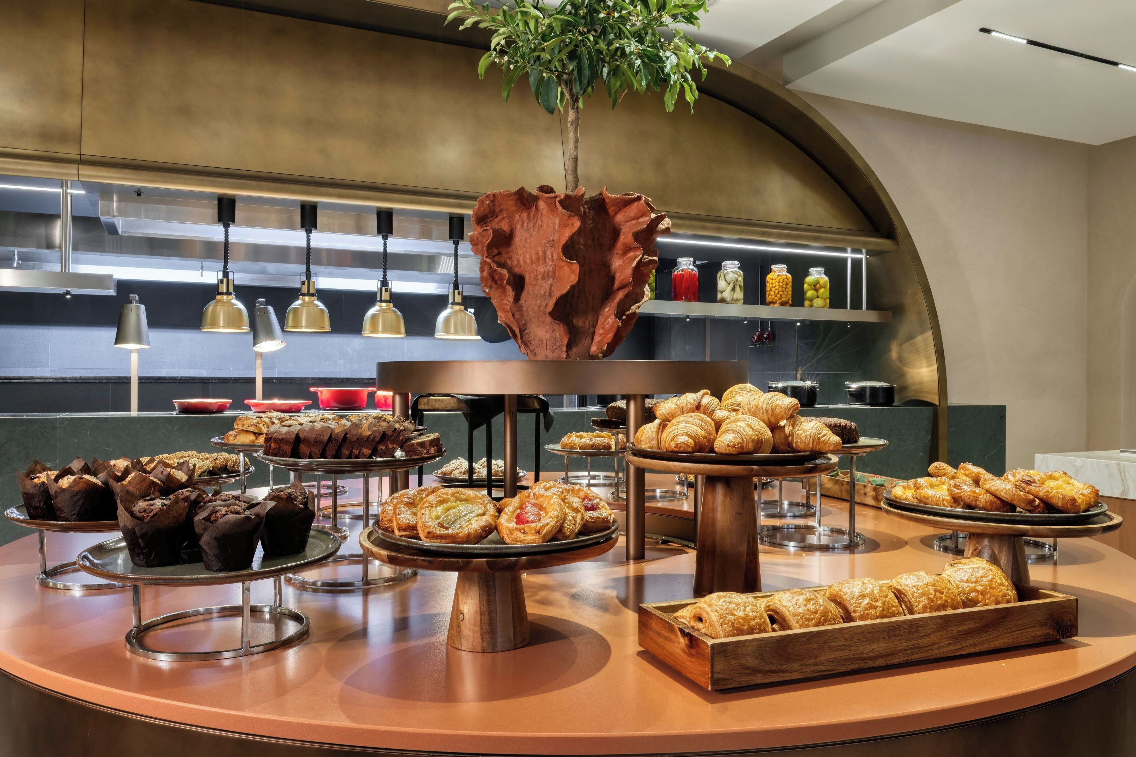Un bufé de desayuno con diversos pasteles y pan, un centro de mesa de madera y una planta colgando del techo.