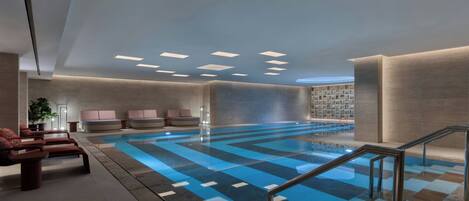 A modern indoor swimming pool with a checkered floor, lounge chairs, and a glass railing.