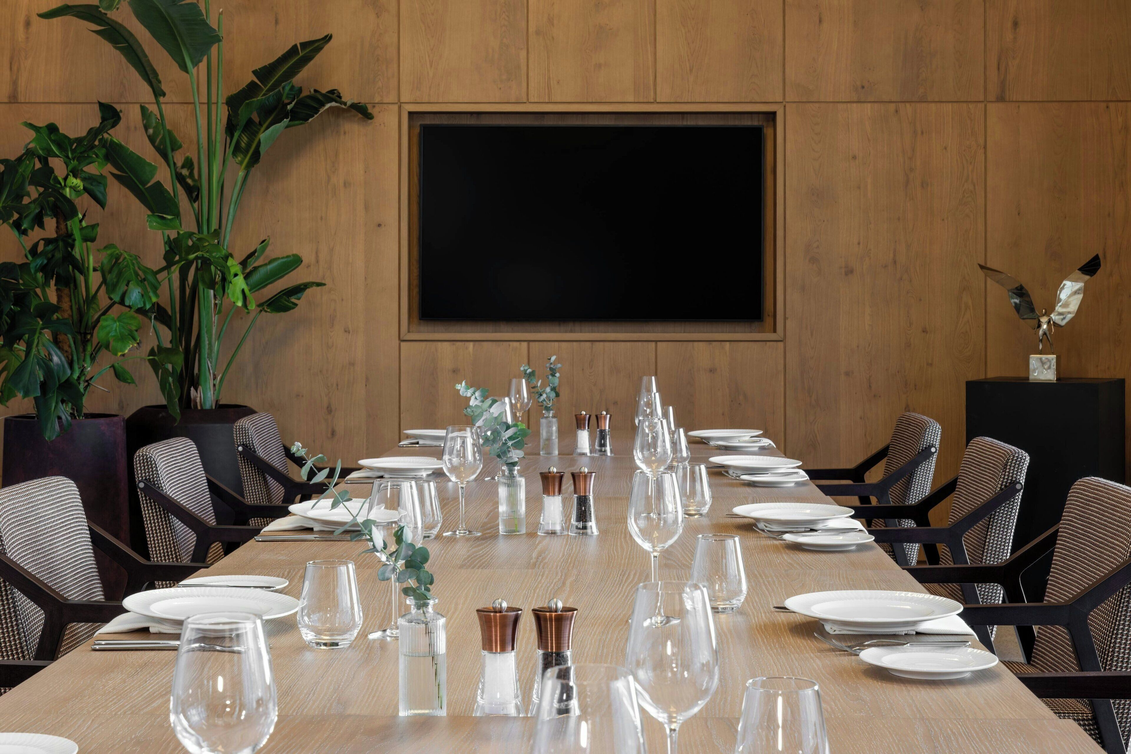 A conference room with a long table set for a meeting, surrounded by chairs, a large screen, and potted plants.