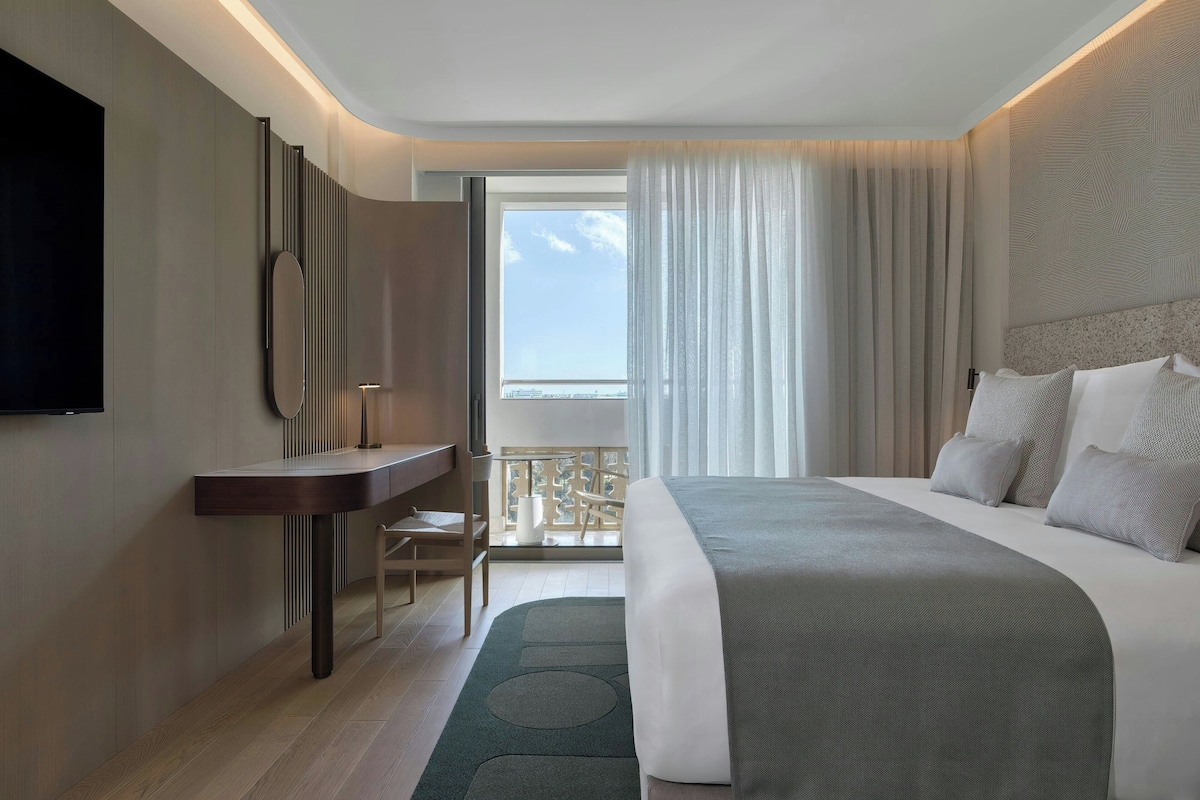 A modern hotel room with a bed, a desk, a chair, a TV, and a balcony with a view.