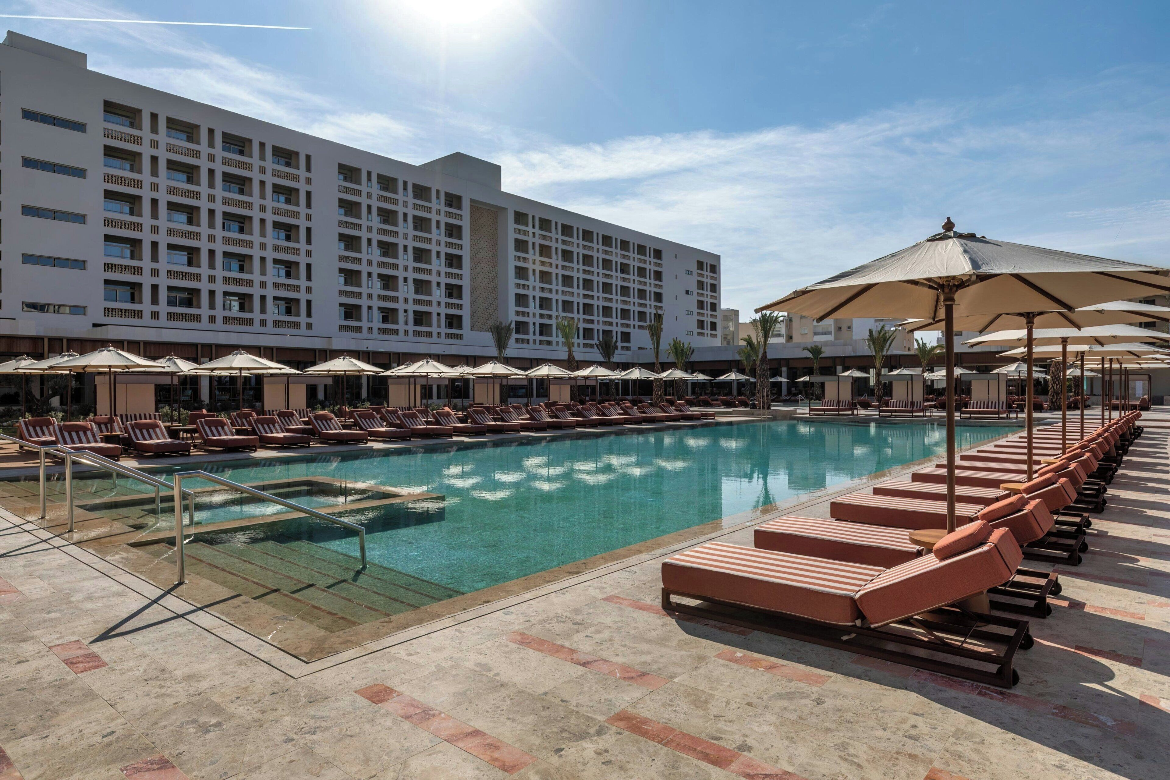 Un espace piscine d’hôtel avec des chaises longues, des parasols et un grand bâtiment en arrière-plan.