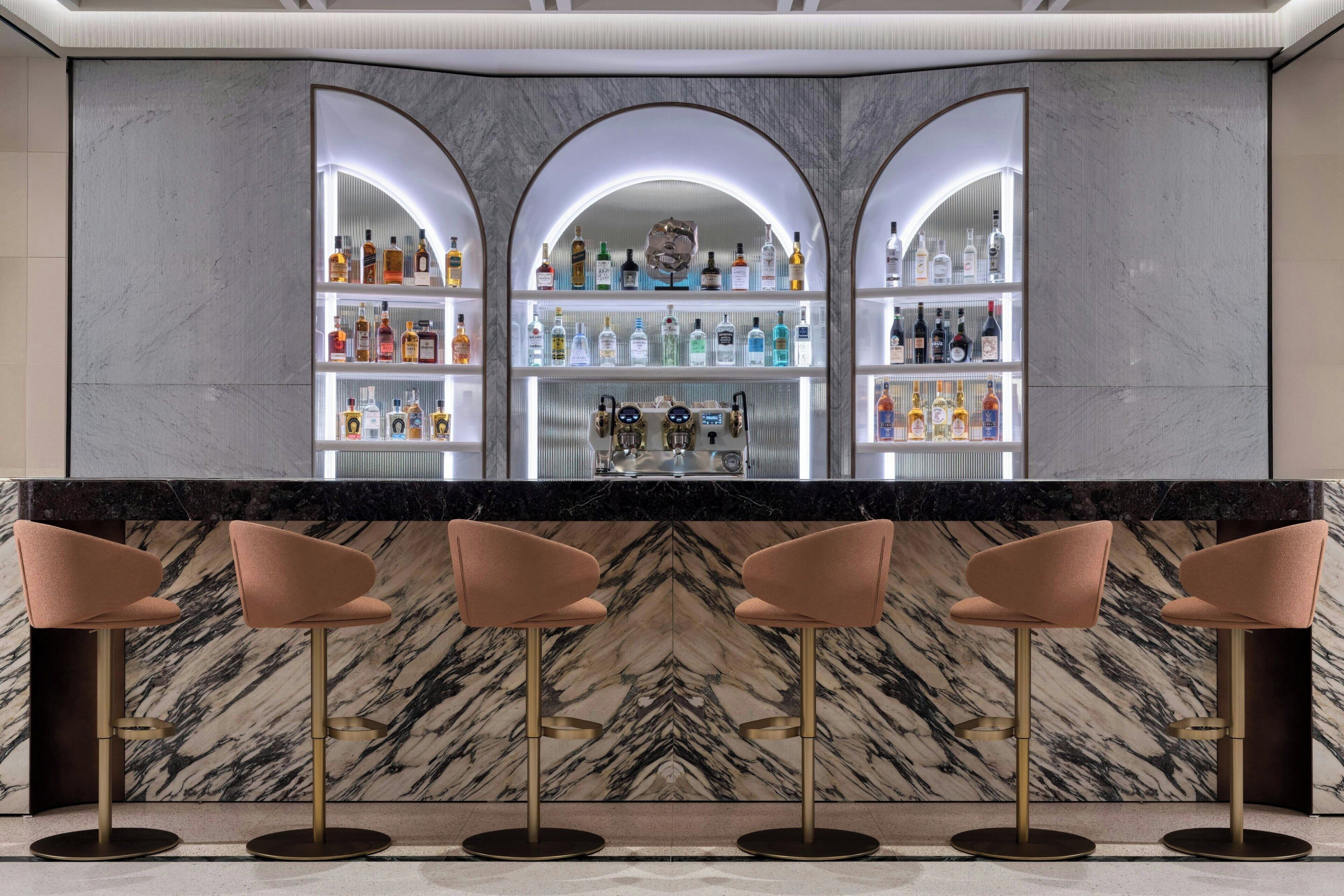 Zona de bar con trasfondos de mármol, una variedad de botellas de licor y taburetes modernos.