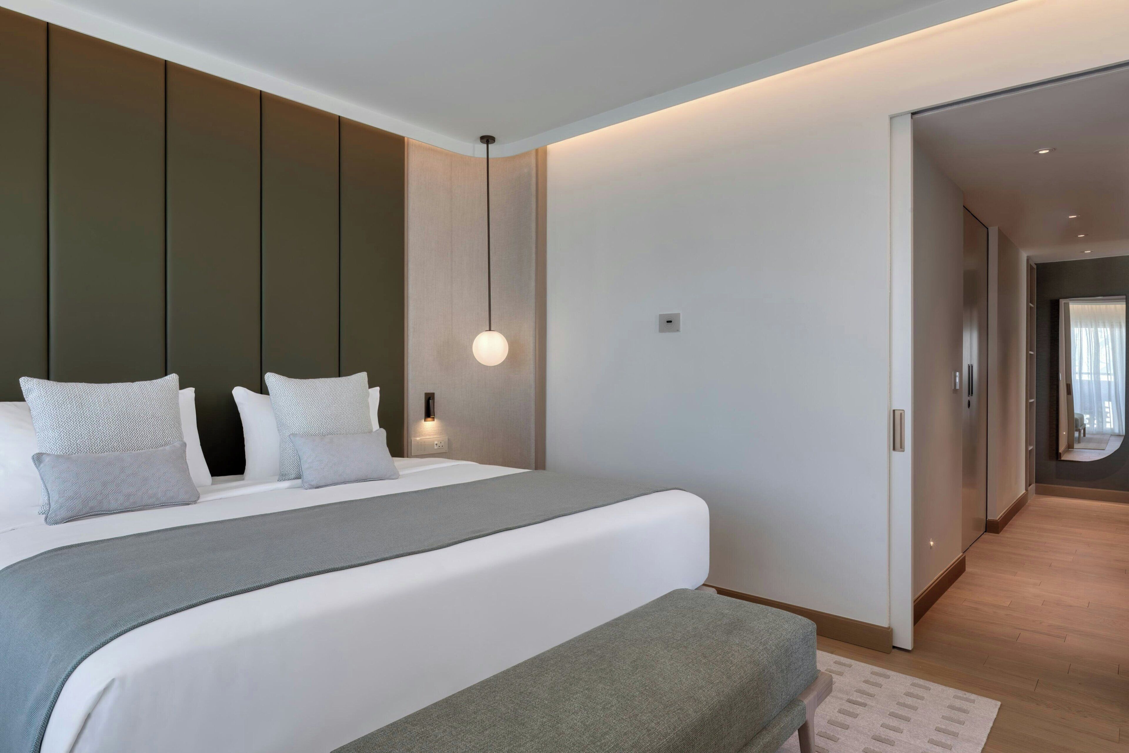 Une chambre d’hôtel moderne avec un grand lit, une table de chevet, un luminaire suspendu et une vue sur le couloir.