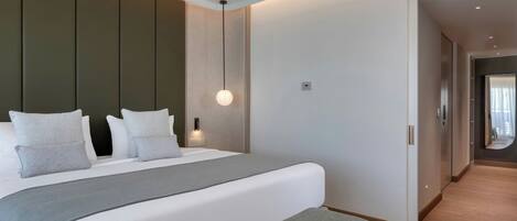 A modern hotel room with a large bed, a bedside table, a hanging light fixture, and a view into the hallway.