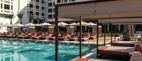 Zona de piscina con sillones de playa y sombrillas, rodeada por un edificio con varios balcones.