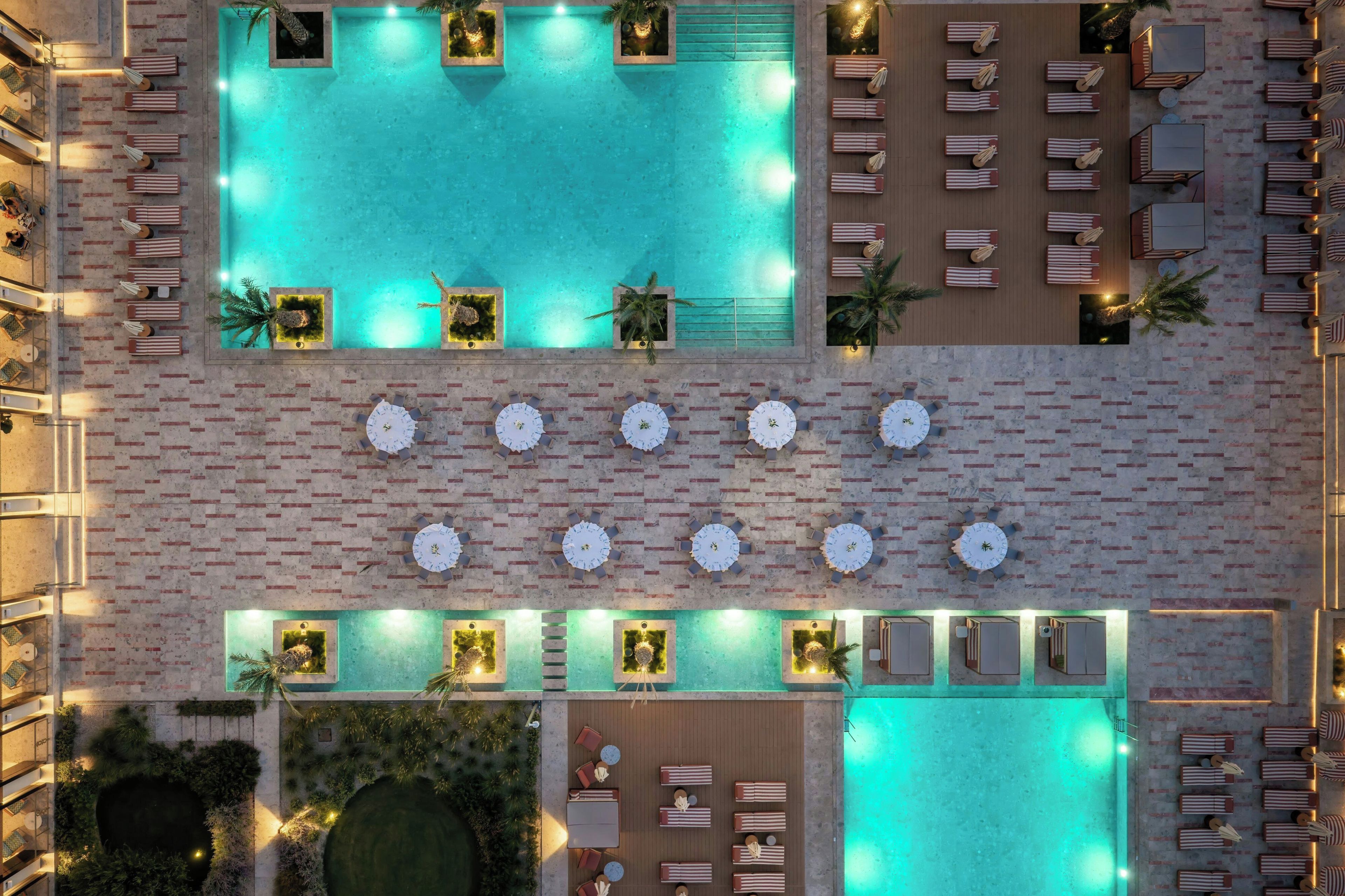 Vista aérea de un área de piscina hotelera con múltiples tumbonas y mesas, rodeada por un muro de ladrillo.