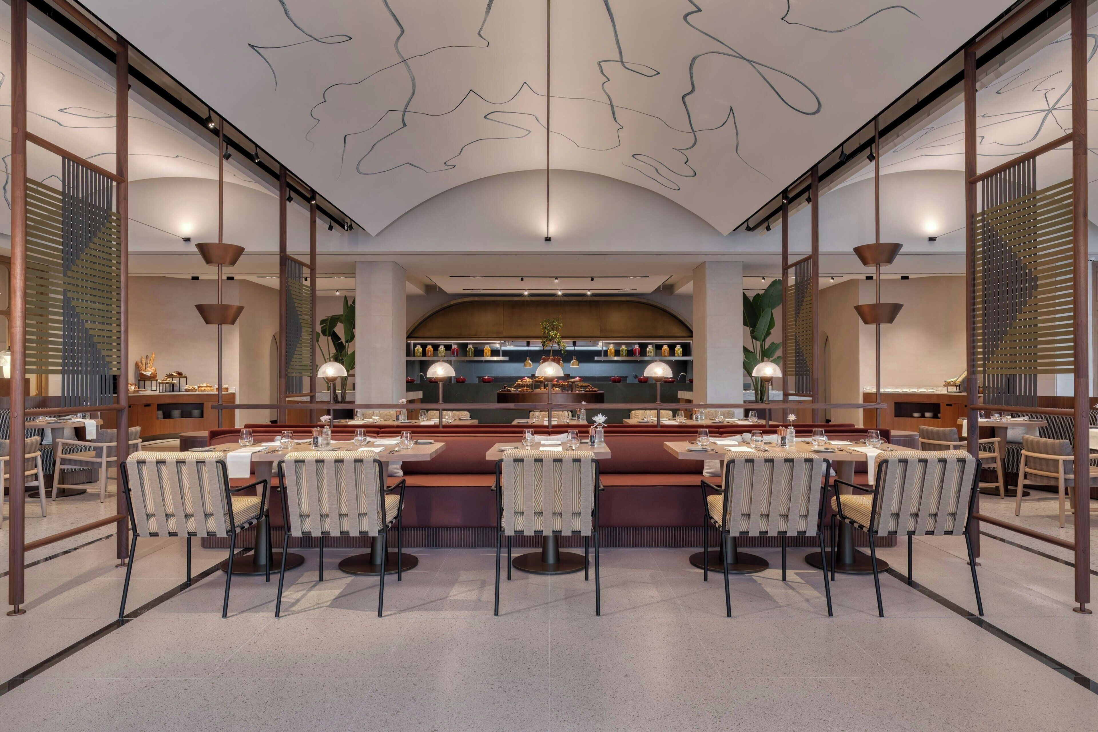 Un restaurante moderno con una barra central, varias mesas de comedor y un techo alto decorado con arte abstracto en líneas.