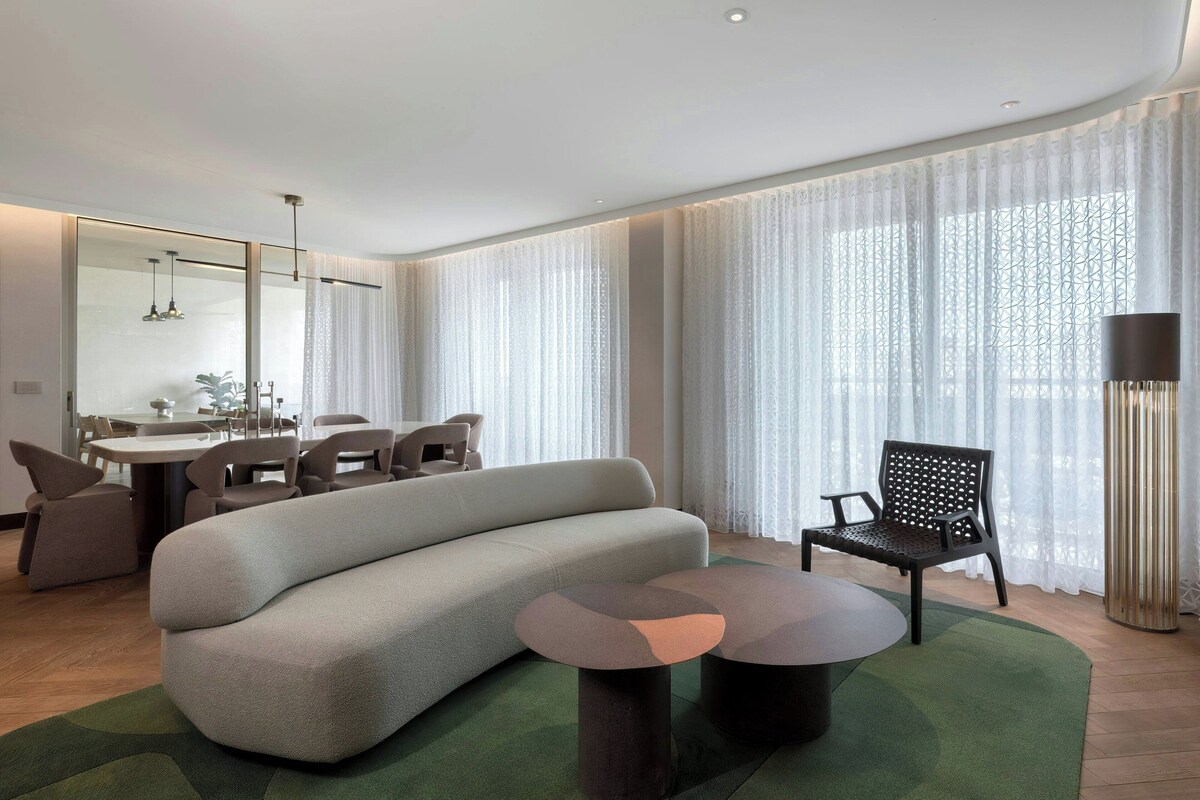 Una sala de estar moderna con un sofá, una mesa de comedor, sillas y un amplio ventanal con cortinas ligeras.