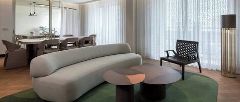 A modern living room with a sofa, a dining table, chairs, and a large window with sheer curtains.