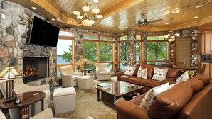 A cozy living room with stone walls, a fireplace, comfortable seating, and large windows offering a view of the outdoors. - Lakefront Torch Lake Lodge  - 4 Floors of Luxury, 8 Bedrooms, Private Dock (Kewadin)