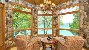 A stone-walled room with large windows overlooking a lake, furnished with patterned chairs and a wooden table. - Lakefront Torch Lake Lodge  - 4 Floors of Luxury, 8 Bedrooms, Private Dock (Kewadin)