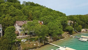 A coastal property with a stone house, dock, and clear turquoise waters. - Lakefront Torch Lake Lodge  - 4 Floors of Luxury, 8 Bedrooms, Private Dock (Kewadin)