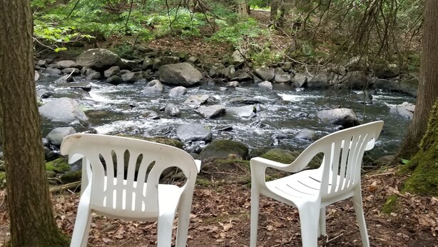 Dos sillas de plástico blancas mirando un arroyo con rocas y árboles al fondo.