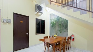 A dining area with a wooden table and chairs, a door with a sign, an air conditioning unit, a window with a view of trees, and a fire extinguisher. - OYO 90037 Rumah Verde (Bogor)
