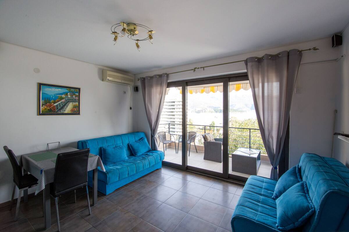A living room with a blue sofa, a dining table with chairs, and a balcony with outdoor furniture.