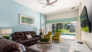 Amplia sala de estar con un sofá de cuero marrón, un sillón amarillo y una mesa de centro. Se observa un ventanal con vistas a una piscina y follaje.