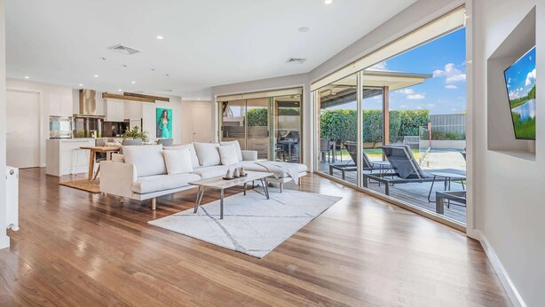 A spacious living area with a large sofa, a coffee table, and a dining table. There is a kitchen area with modern appliances and a large window overlooking a garden. - 6A Blair St - Echuca Holiday Homes (Moama)