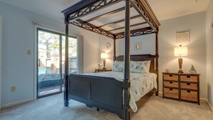 A four-poster bed with a dark finish, a nightstand with a lamp, and a decorative wall piece. - Sound side OBX Condo no stairs, pool, enjoy the Fall, less crowds peaceful ! (Kitty Hawk)