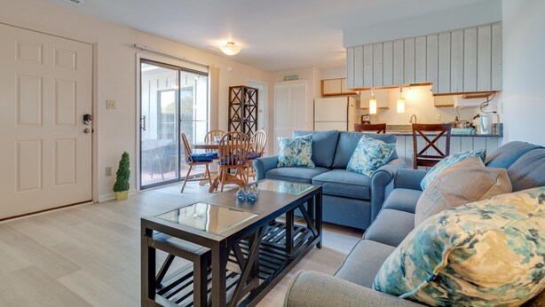 A modern living room with a glass coffee table, blue sofas, and a dining area with wooden chairs. - Sound side OBX Condo no stairs, pool, enjoy the Fall, less crowds peaceful ! (Kitty Hawk)