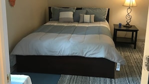 Een net opgemaakt bed met een gestreept dekbed en meerdere kussens, een nachtkastje met een lamp, en een klein decoratief item aan de muur.