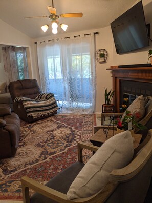 Uma sala de estar com lareira, ventilador de teto e uma janela grande com cortinas leves.