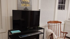 A living room with a black entertainment unit holding a flat-screen TV, a book, and a framed picture. There's a wooden rocking chair with a blanket. A poster on the wall reads "LIVE. TRAVEL. EXPLORE." - Cozy So End Suite (Burlington)