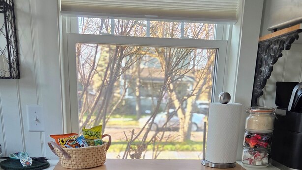 A kitchen with a stainless steel refrigerator, a wooden countertop, a basket of snacks, a jar of sugar, and a window with a view of trees and a car. - Cozy So End Suite (Burlington)