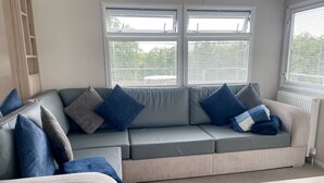 Una sala moderna con un sofá seccional gris, varios cojines, una mesa de centro de madera y ventanas amplias con persianas.