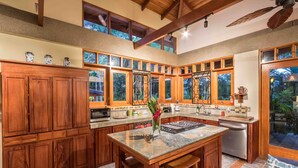 A spacious kitchen with wooden cabinets, a central island, and large windows offering views of the outdoors. - Sonidos Del Mar - Tropical Garden Villa (Jaco)