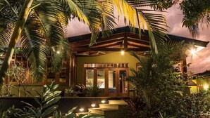 A modern house with a wooden exterior, a glass door entrance, and a well-lit garden area with various plants. - Sonidos Del Mar - Tropical Garden Villa (Jaco)