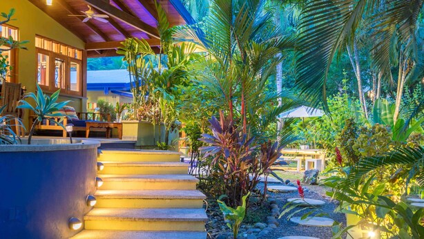 A tropical garden with stone steps leading to a wooden deck, surrounded by lush greenery and palm trees. - Sonidos Del Mar - Tropical Garden Villa (Jaco)