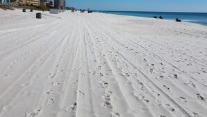 Ein weiter, sandiger Strand mit Fußspuren, die zum Meer führen, Gebäude im Hintergrund und ein klarer, blauer Himmel.
