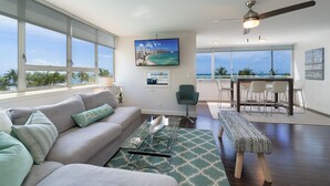 Un vaste espace de vie avec une grande fenêtre donnant sur une plage, un ventilateur de plafond et un coin repas avec des chaises.