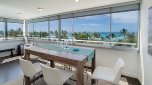 Un espace repas avec une table en bois, des chaises blanches et une vue sur l’océan grâce à de grandes fenêtres.