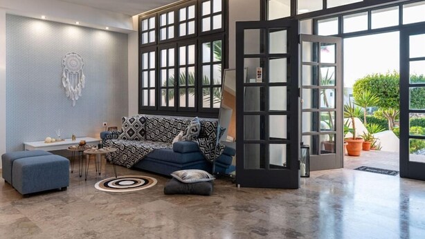 A modern living room with a blue sofa, a dreamcatcher wall art, and a large window with a view of greenery. - Anemos Inn Adults Only By Estia (Hersonissos)