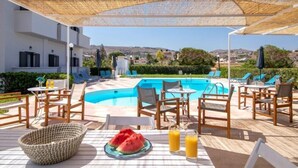 A poolside area with lounge chairs, a table with a plate of watermelon and drinks, and a view of the surrounding landscape. - Anemos Inn Adults Only By Estia (Hersonissos)