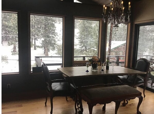 A dining area with a large table, chairs, and a bench. There are three large windows with a view of snow-covered trees.
