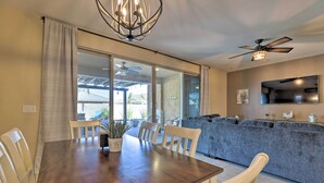 Un coin repas avec une table en bois et des chaises blanches, un ventilateur de plafond et une grande fenêtre donnant sur l’extérieur.