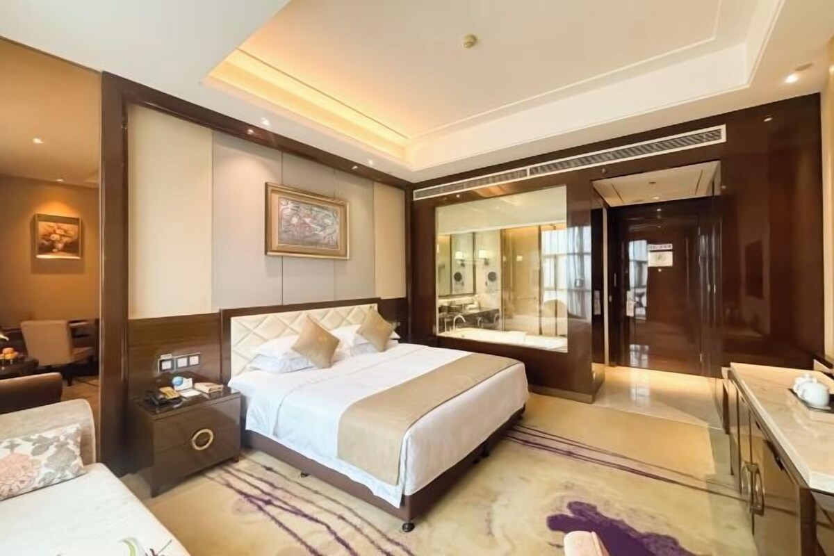 Habitación de hotel con una cama grande, una mesita de noche, un escritorio, un espejo y un baño visible a través de una puerta de cristal.