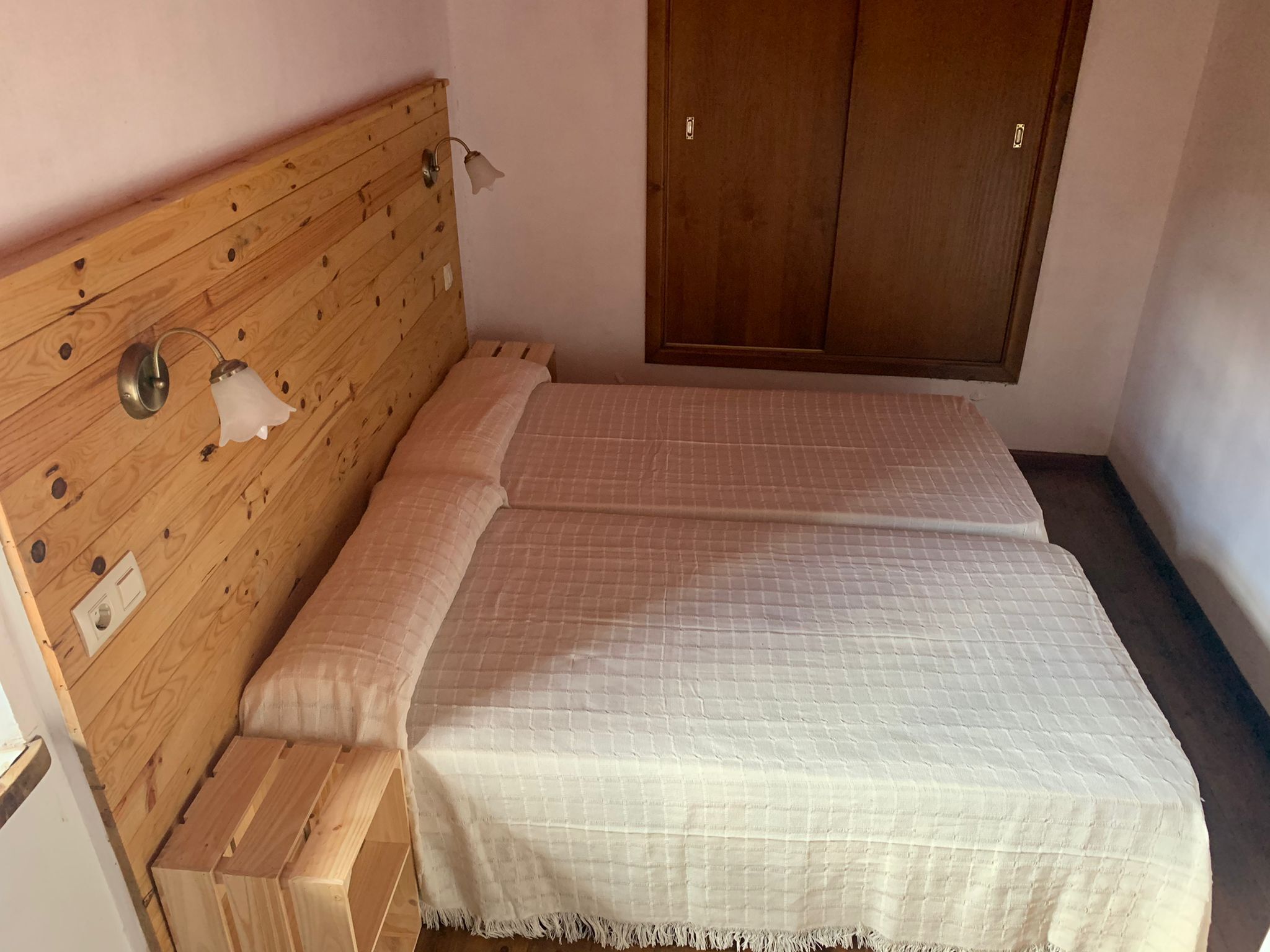Huoneessa on puinen sängynpääty, valkoinen, kuviollinen peitto sängyllä, puinen vaatekaappi ja seinään kiinnitetty valaisin.