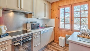 A modern kitchen with stainless steel appliances, wooden cabinets, and a large window with a view of trees. - Le Chalet des Neiges - Welkeys (Megève)